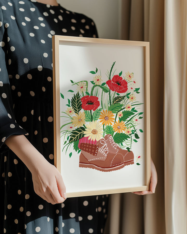 Person in a polka dot dress holding a framed botanical Art Print titled 'Wildflower Walk' showcasing a whimsical bouquet of wildflowers sprouting from a pair of rustic boots.