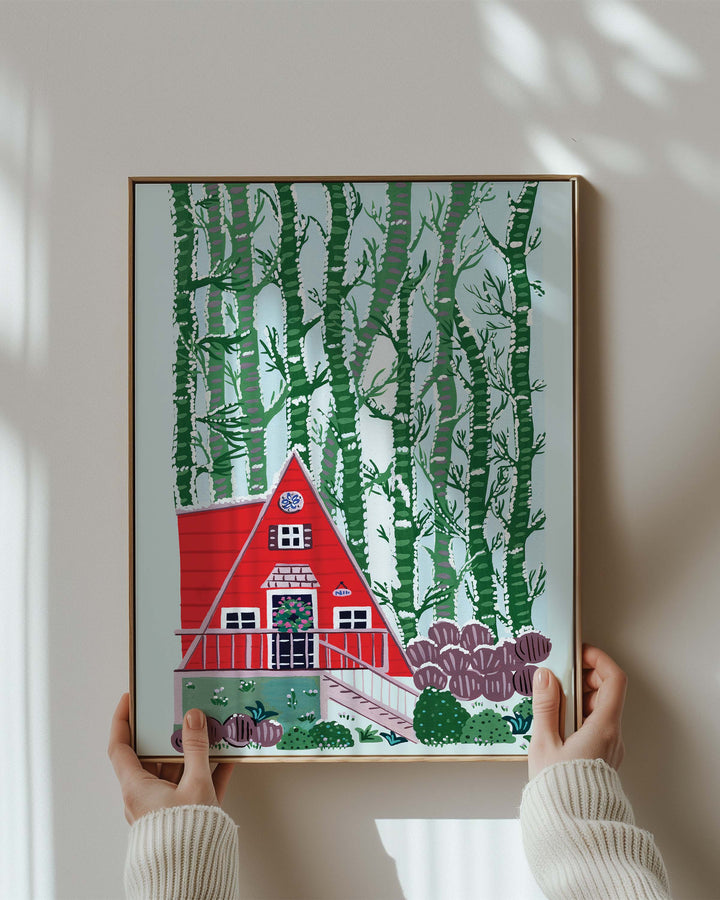 Person holding a Framed Artwork "Woodland Hideaway" features a charming red A-frame cabin nestled amidst a dense stand of birch trees.