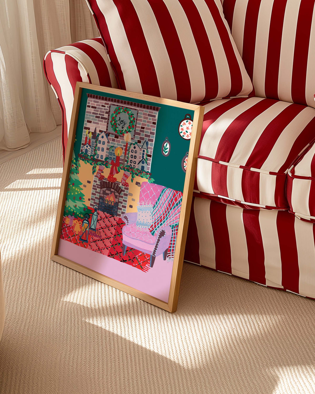 Framed Art Print titled 'Yuletide Haven' by Little Berries Studio, propped against a red and white striped sofa in a bright, sunlit room.